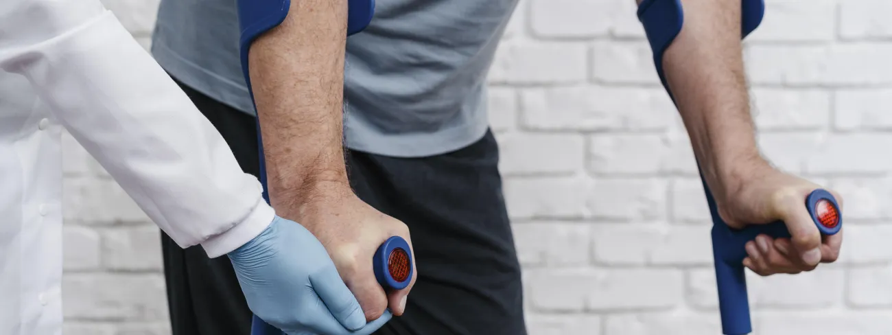 Mężczyzna z kulami, asystowany przez osobę w białym kitlu i rękawiczkach.